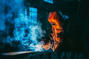 A welder in a workshop using safety gear to weld metal, surrounded by blue smoke and atmospheric lighting.
